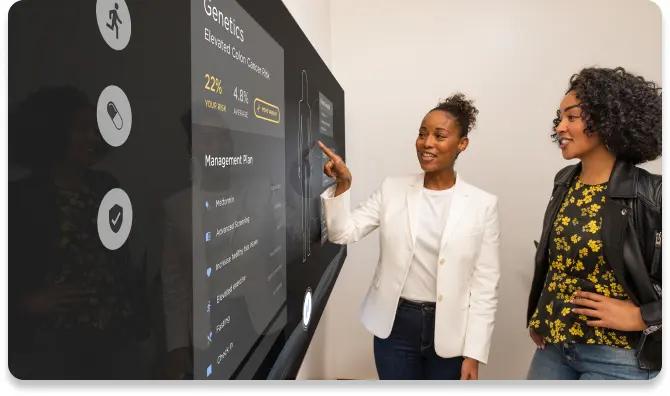 Young female patient looking at a female doctor in an exam room with the Smart Screen showing her health information in the background.