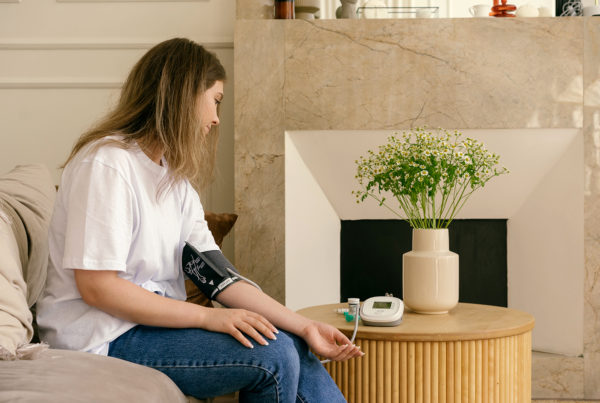 young woman sits on couch getting her blood pressure taken with a machine on her side table